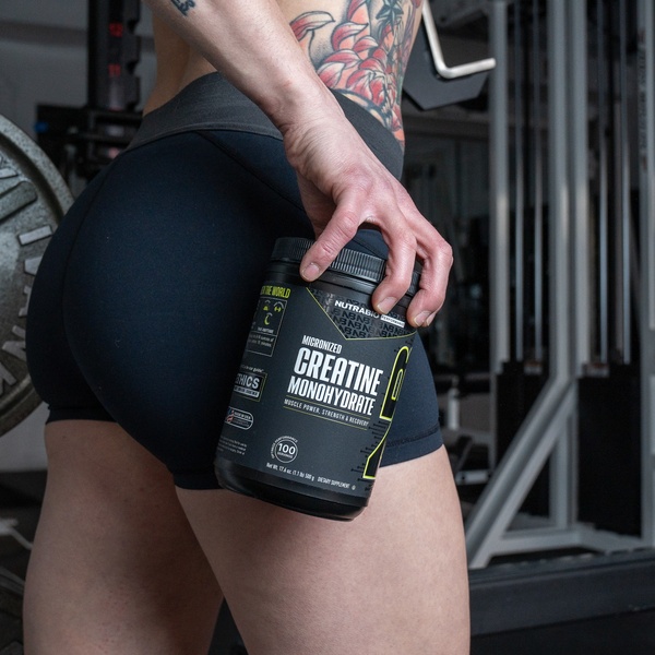A close-up photograph of a user holding a Creatine Monohydrate supplement tub inside a gym environment to establish brand authority and product utility.